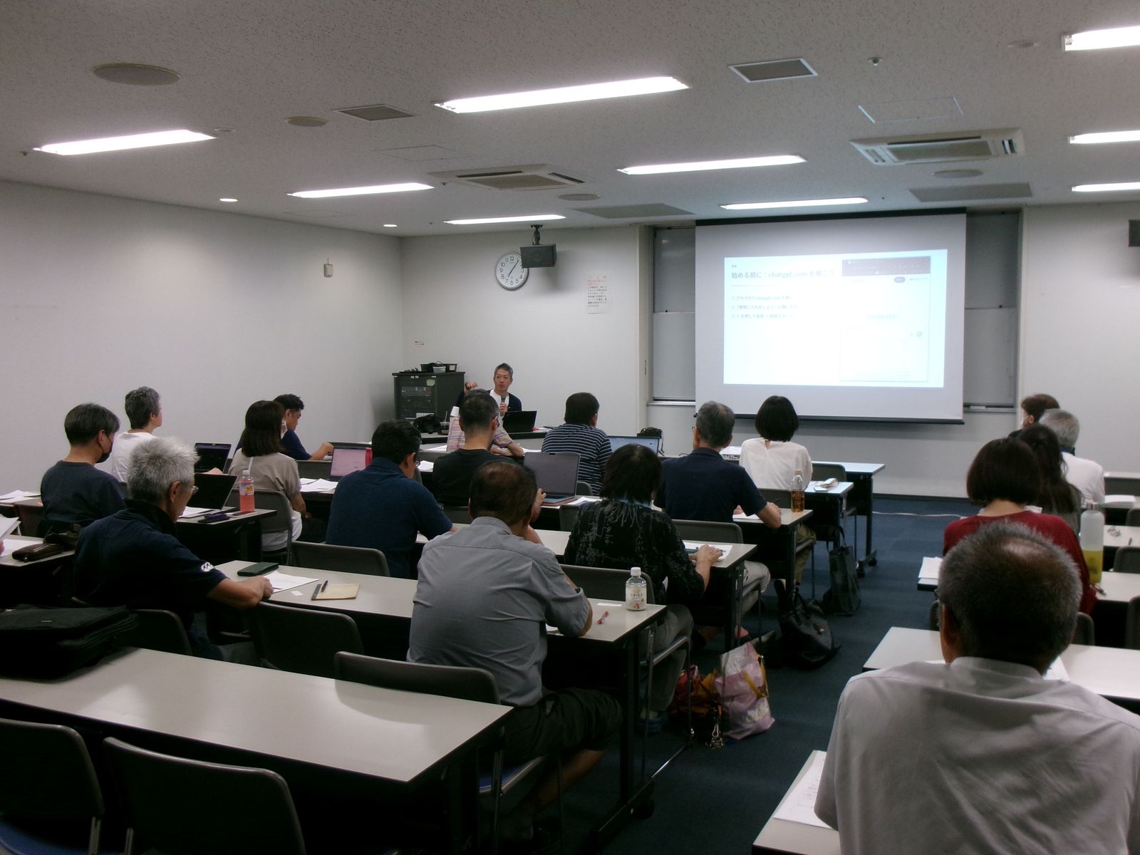 福井市中央公民館ICT講座「AI活用術」の会場。アオッサ6階の研修室に教室形式で30名が着席し、スクリーンには「始める前に：chatgpt.com を開こう」のスライドが投影されている